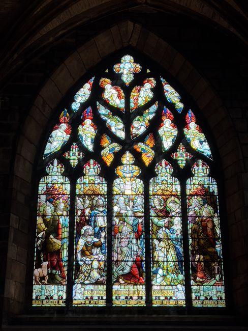 A large, colorful stained glass window featuring intricate biblical and religious motifs, set within a dark stone arch framing. The window depicts multiple figures, some holding objects such as scrolls and staffs, with decorative geometric patterns and floral designs at the top. The surrounding area shows a dimly lit interior, suggesting the window is part of a church or cathedral, and illuminating the interior with vibrant light. The scene aligns with the context of historic ecclesiastical buildings, which may require careful packing and handling during house removals or relocations, as carried out by services like Man with Van St Giles.
