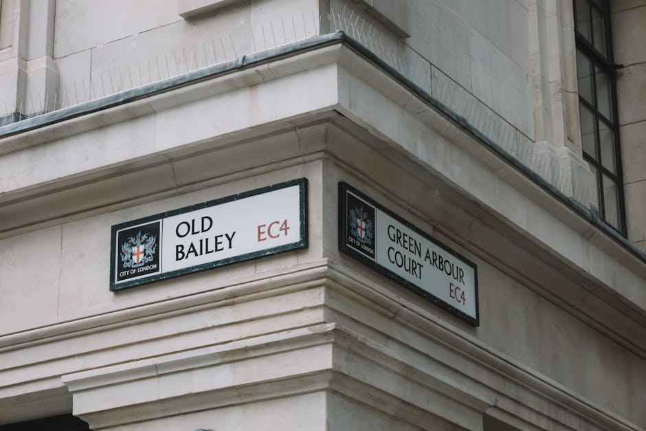 Photograph of the corner of a beige stone building with two street signs attached to the facade. The first sign reads 'OLD BAILEY EC4' and features a logo with a crown and a shield, indicating it is part of the City of London, while the second sign states 'GREEN ARBOUR COURT EC4' with the same logo. The signs are affixed just below a balustrade and above a window with black frames. The building appears to be part of a historic or official structure, with a tidy appearance and minimal exterior decorations. The context suggests a street within central London, relevant to residential or business relocations, where careful furniture transport and packing may be required during house removals or moving processes.
