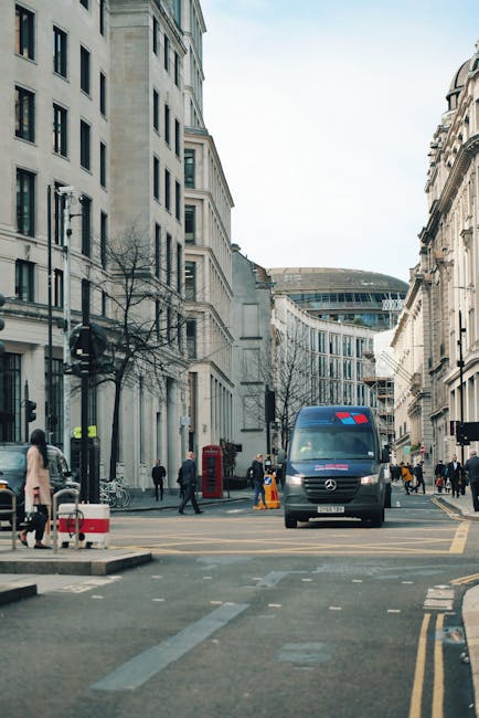 A street scene in central London with multi-storey, light-colored stone buildings lining the road, some with decorative architectural features. A black Mercedes-Benz van with a roof-mounted blue light is parked on the road, part of a home relocation or furniture transport process, supported by Man with Van St Giles. Several pedestrians walk along the pavement and cross the street, including individuals in casual and business attire. A red British telephone booth and a black post box are visible on the sidewalk, along with leafless trees suggesting late autumn or winter. The street is marked with yellow double lines, and orange traffic cones are placed near the van, indicating ongoing loading or unloading activities. The lighting is natural daylight, and the scene depicts a typical busy London intersection with active moving and packing logistics occurring as part of a professional removals service.