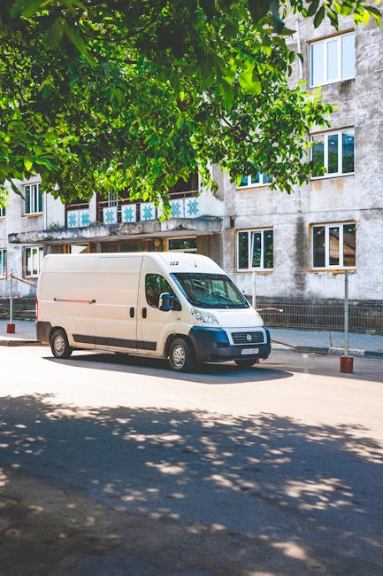 A white cargo van parked on a residential street with multi-storey apartment buildings in the background. The van is positioned parallel to the pavement, with the driver's side facing outward, and app