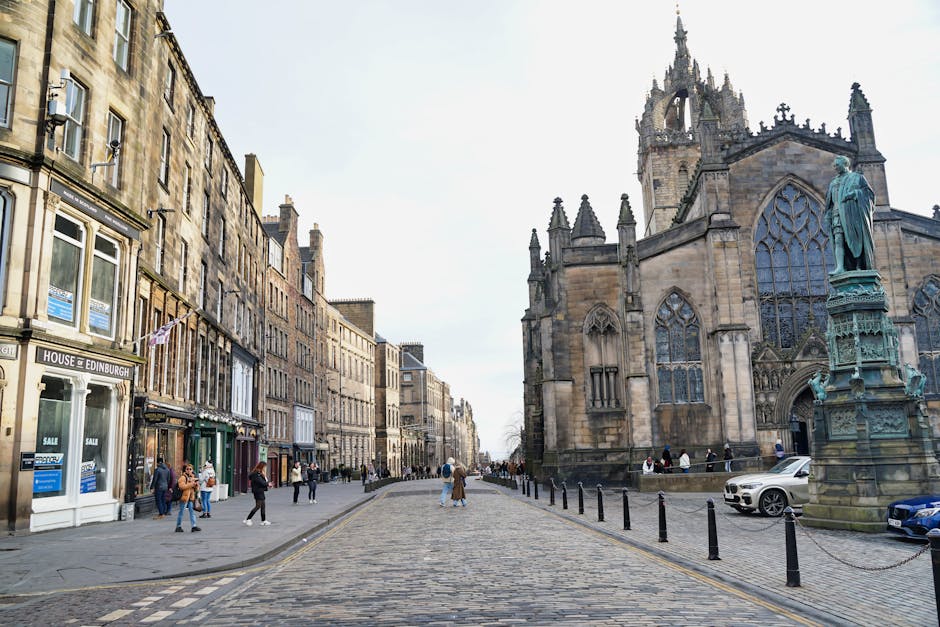 A wide cobbled street in a historic city center featuring a large Gothic-style stone church with intricate stained-glass windows and a tall spire, alongside traditional sandstone buildings with large display windows, some advertising property sales. Pedestrians are walking along the pavement, and an assortment of cardboard boxes and wrapped furniture is visible near the church doorway, suggesting recent home or office move activities. A silver and a dark blue vehicle are parked adjacent to the church, with bollards lining the edge of the street to separate pedestrian areas from parked vehicles. The scene is outdoors during daylight with clear weather, and a moving service provider such as Man with Van St Giles may be involved in home relocation or furniture transport, highlighted by the presence of packed boxes and packing materials within the scene.