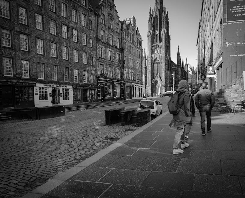 A wide cobbled street in a historic city center featuring a large Gothic-style stone church with intricate stained-glass windows and a tall spire, alongside traditional sandstone buildings with large display windows, some advertising property sales. Pedestrians are walking along the pavement, and an assortment of cardboard boxes and wrapped furniture is visible near the church doorway, suggesting recent home or office move activities. A silver and a dark blue vehicle are parked adjacent to the church, with bollards lining the edge of the street to separate pedestrian areas from parked vehicles. The scene is outdoors during daylight with clear weather, and a moving service provider such as Man with Van St Giles may be involved in home relocation or furniture transport, highlighted by the presence of packed boxes and packing materials within the scene.