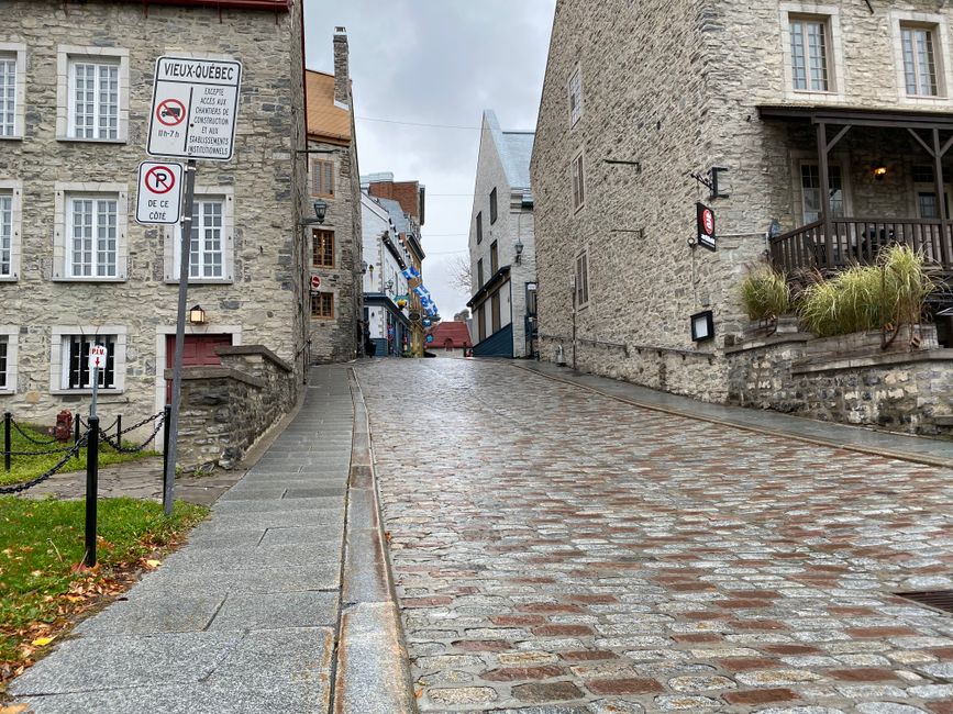 A cobbled uphill street in St Giles, lined with historic stone buildings featuring multiple windows and traditional facades. On the left side, there is a sidewalk bordered by a small grassy area with a black chain fence. A signpost indicates local restrictions for construction and parking within the area. On the right, a building with a balcony and potted plants extends over the pavement. The scene appears wet, suggesting recent rain, and the overcast sky creates diffused lighting. In the context of house removals and furniture transport, this setting may illustrate a typical street where loading and unloading operations take place, with Man with Van St Giles potentially managing home relocation activities in such environments.