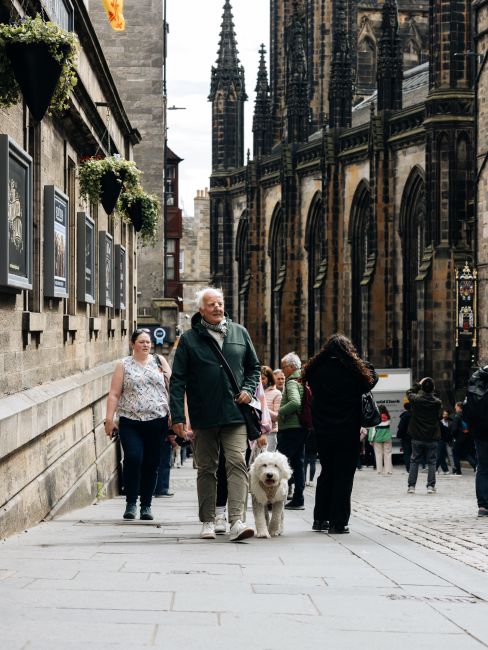 A wide view of a cobbled street in St Giles, with historic stone buildings lining both sides and a large, Gothic-style church featuring intricate stained glass windows and towering spires on the right. In the foreground, a group of pedestrians is walking along the pavement, some carrying shopping bags or backpacks, while others stand near the sidewalk. Several parked vehicles, including a white SUV, are positioned along the street and on the pavement near the church, with black bollards separating the parking area from the pedestrian zone. In the background, the street extends into the distance under an overcast sky, with additional pedestrians, street furniture, and shopfronts visible, illustrating a busy urban environment suitable for local home removals and furniture transport, supported by services like those offered by Man with Van St Giles for packing and moving logistics.