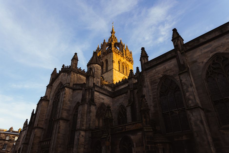 Photograph of an exterior view of a historic Gothic-style church with large stained glass windows featuring intricate tracery. The church's façade is constructed from grey stone with elaborate archit