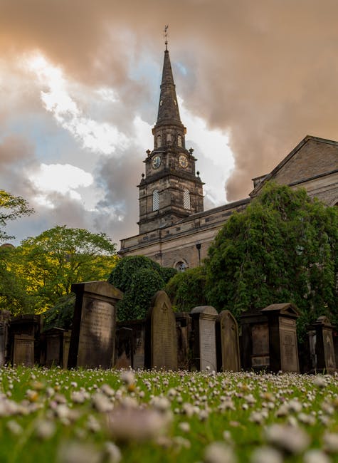 A tall church steeple with a clock face extending into a cloudy sky, situated behind a graveyard with numerous headstones of various shapes and sizes. Surrounding the graveyard are lush green trees and bushes, partially obscuring the building's stone facade. The scene depicts an outdoor setting typical of a historic church or cathedral located in St. Giles, with natural lighting highlighting the stonework and greenery. This environment reflects a peaceful, well-maintained area likely involved in relocating or moving services, such as those offered by Man with Van St Giles, with a focus on careful handling of items within or around such historic sites.