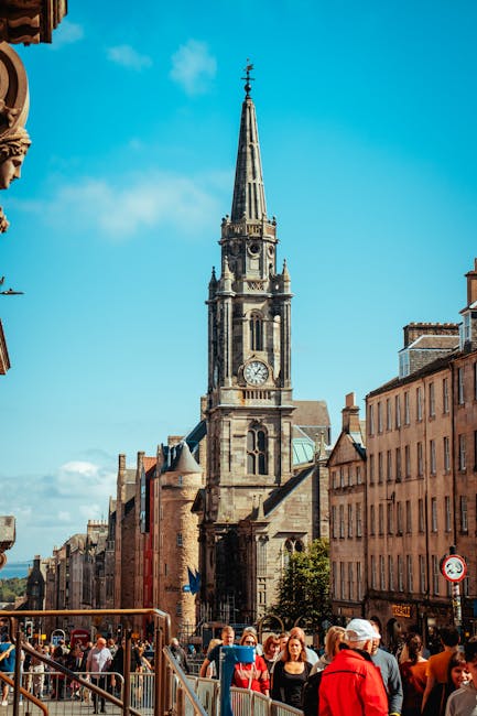 A tall church steeple with a clock face extending into a cloudy sky, situated behind a graveyard with numerous headstones of various shapes and sizes. Surrounding the graveyard are lush green trees and bushes, partially obscuring the building's stone facade. The scene depicts an outdoor setting typical of a historic church or cathedral located in St. Giles, with natural lighting highlighting the stonework and greenery. This environment reflects a peaceful, well-maintained area likely involved in relocating or moving services, such as those offered by Man with Van St Giles, with a focus on careful handling of items within or around such historic sites.