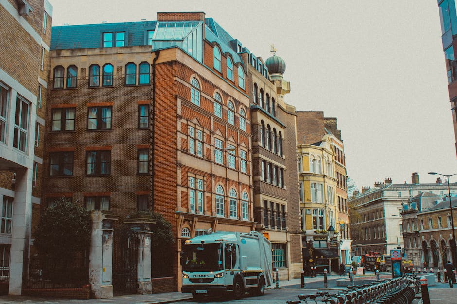 A street scene near St Giles Circus showing several multi-storey buildings with a variety of architectural styles, including brick and ornate facades, some with bay windows and decorative elements. In the foreground, a light blue van belonging to Man with Van St Giles is parked on the street, positioned near loading bays used for home relocation and furniture transport services. The van is adjacent to the pavement, beside a row of bike racks, with some movement of boxed and wrapped furniture visible, suggesting ongoing packing and loading for a house removal. There are a few pedestrians and street signage present, with cloudy daylight illuminating the scene, reflecting typical urban loading and loading area conditions as part of a professional moving process involved in house removals and relocation services.