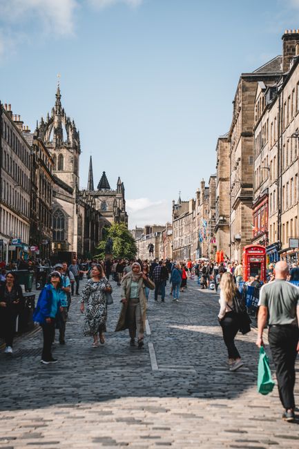 A busy street scene in St Giles with a mix of pedestrians walking along a cobblestone road, some carrying shopping bags, and others engaged in conversation. On the right side, a person in casual clothing is pushing a hand truck loaded with cardboard boxes and wrapped furniture pieces toward a white van parked near the sidewalk, likely involved in a home relocation or furniture transport process. The background features historic stone buildings, and a church with gothic architecture is visible beneath a clear blue sky. Overhead, the environment is well-lit with natural daylight, and the surrounding scene indicates a typical urban area suitable for household moving services. Man with Van St Giles occasionally appears as part of the scene, emphasizing professional removals in a city setting.