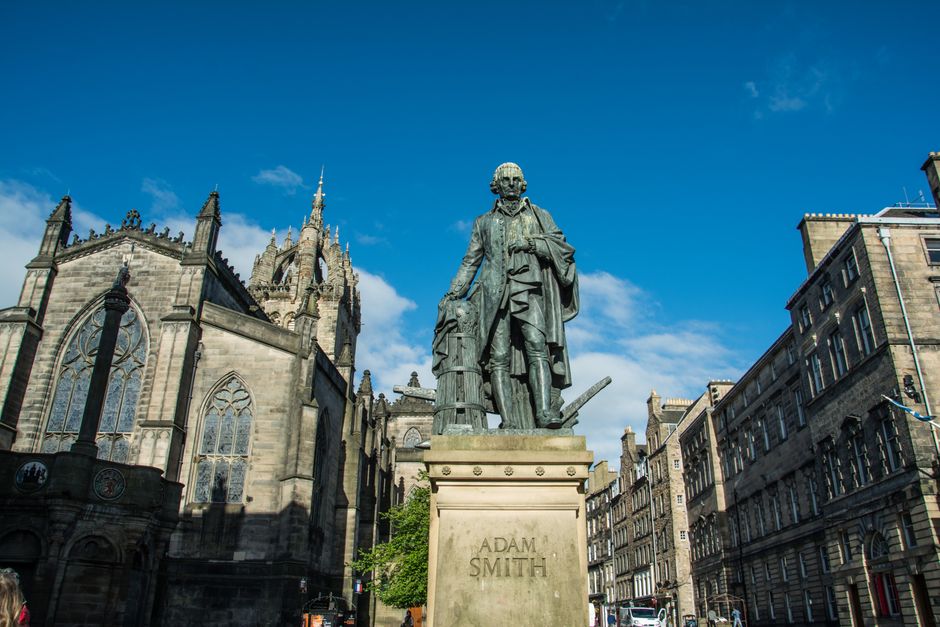A bronze statue of Adam Smith standing on a stone pedestal, located in an outdoor urban setting. The statue depicts Smith dressed in period clothing, with one hand resting on a book and the other holding a scroll. The surroundings include historic stone buildings with ornate Gothic-style architecture and stained glass windows, as well as other classical buildings lining the street. The scene is illuminated by natural daylight, with a clear blue sky and some scattered clouds overhead. In the background, there is a large church or cathedral with pointed spires, and the area appears to be a well-preserved historic part of the city, where house removals, packing, and furniture transport services by Man with Van St Giles may be relevant for home relocation or moving logistics.
